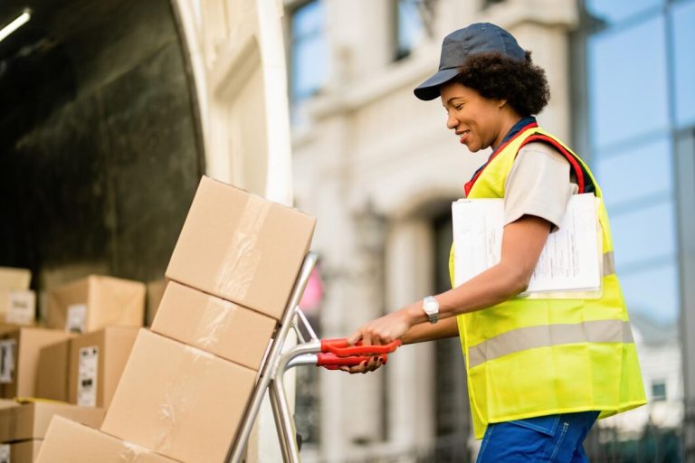 Heureux livreur afro-américain avec camion à main déchargeant des colis d'une camionnette